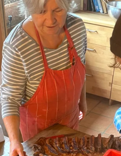 Eine Frau in einer roten Schürze schneidet ein großes Stück geräuchertes Fleisch auf einem Holzbrett in einer Küche.