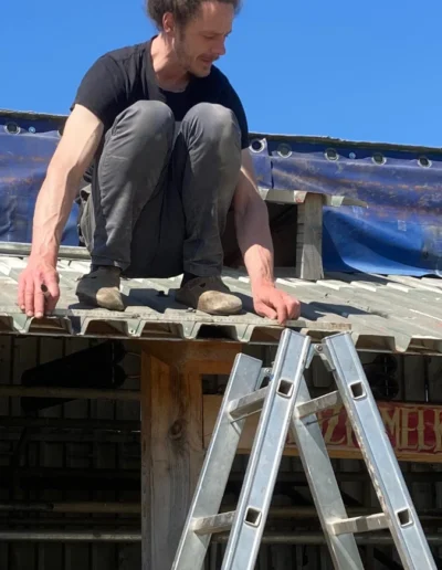 Mann mit Dutt bei Reparaturarbeiten auf einem Wellblechdach, eine Leiter steht im Vordergrund.