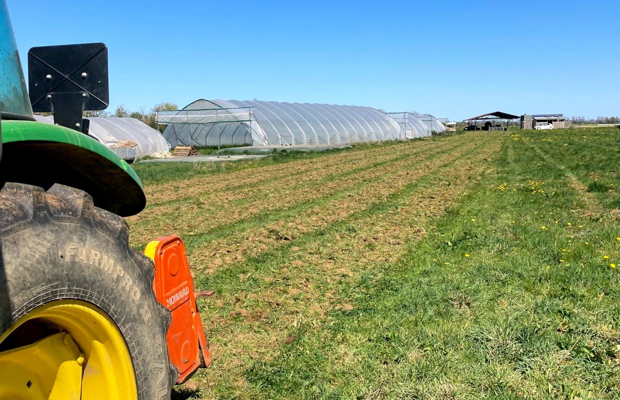Traktor auf dem Acker vor Gewächshäusern Blick von einem grünen Traktor auf ein frisch gepflügtes Feld mit Gewächshäusern im Hintergrund an einem sonnigen Tag auf einem landwirtschaftlichen Betrieb.