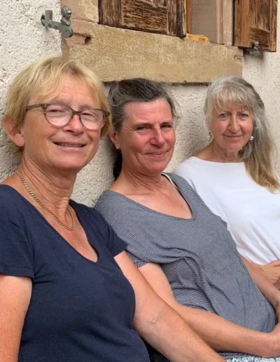 Drei lächelnde Frauen mittleren Alters vom Solawi-Team sitzen entspannt nebeneinander vor einer Hauswand.