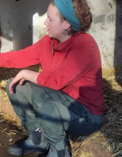 Eine junge Frau aus dem Hof-Team hockt in Arbeitskleidung im Stroh eines Stalls.