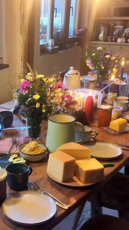 Gemeinschaftliches Essen am reich gedeckten Tisch Reich gedeckter Holztisch in einem rustikalen Raum mit Käse, Aufstrich, Blumensträußen und Kerzenlicht, der eine gemütliche Gemeinschaftsatmosphäre schafft.