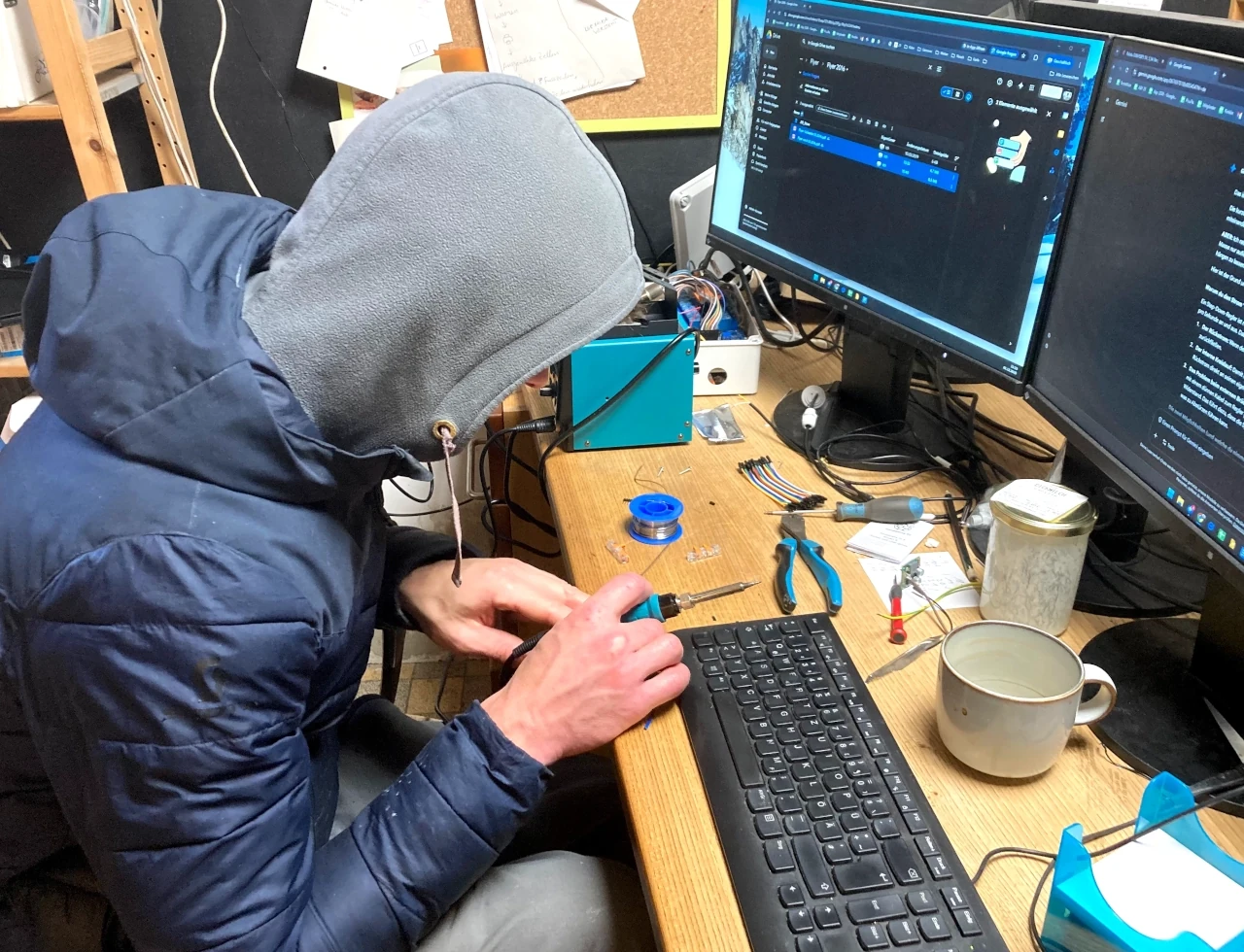 Technische Entwicklung und Reparatur in der Hof-Werkstatt Person mit Kapuze lötet an einem Elektronik-Bauteil an einem Schreibtisch vor zwei Monitoren in einer Werkstatt.