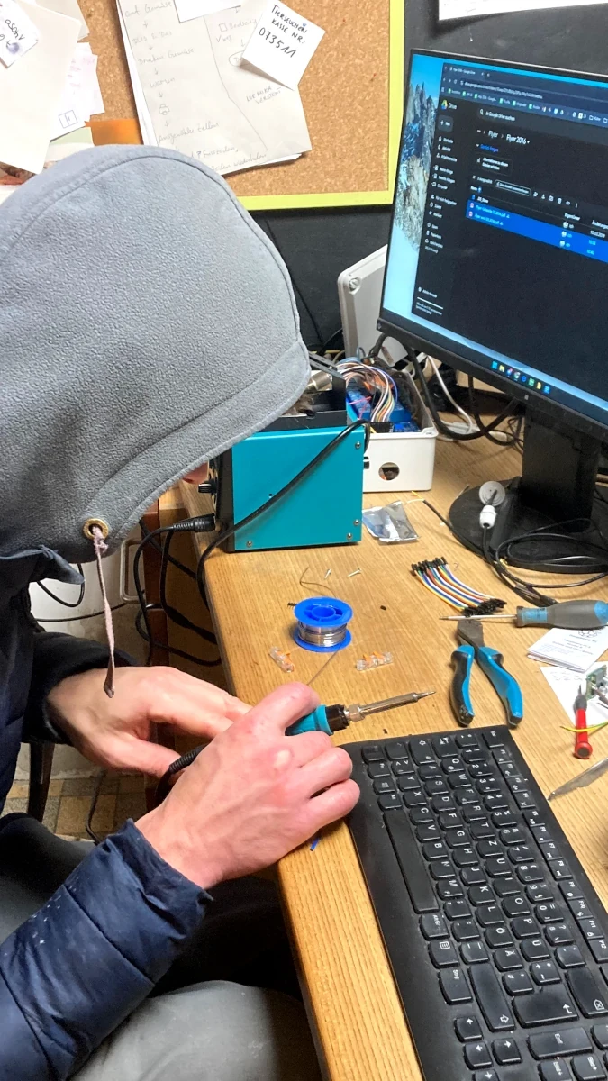 Lötarbeiten in der Hof-Werkstatt Person im grauen Hoodie lötet an einem Holztisch an einer elektronischen Schaltung, umgeben von Werkzeug und einem Computer.