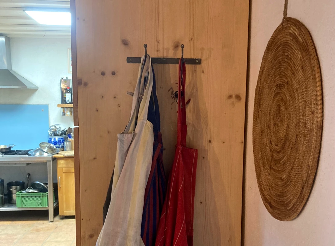 Hofküche mit Schürzen und Taschen an Holzwand Stofftaschen und Schürzen hängen an Haken an einer Holzwand, mit Blick in eine Hofküche im Hintergrund.