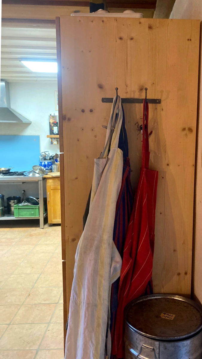 Schürzen an der Wand der Hofküche Bunte Kochschürzen hängen an Haken an einer Holzwand, mit einem Einblick in die betriebsame Hofküche im Hintergrund.