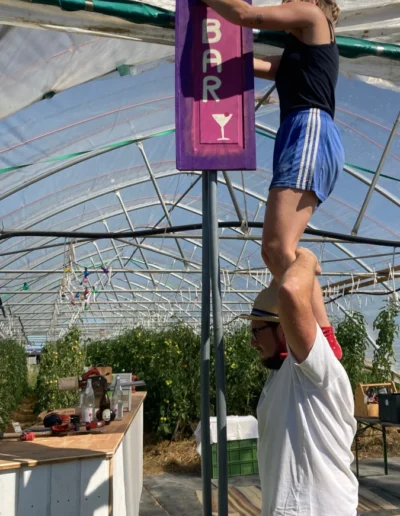 Eine Frau sitzt auf den Schultern eines Mannes in einem Gewächshaus und befestigt ein lila 'BAR'-Schild an einer Stange, im Hintergrund Tomatenpflanzen.