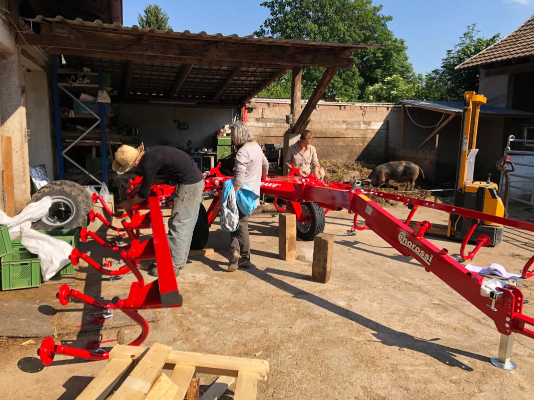 Drei Personen vom Hof-Team bauen gemeinsam einen neuen, roten Heuwender der Marke Enorossi auf dem Hof zusammen. Im Hintergrund ist ein Schwein im Stall zu sehen.