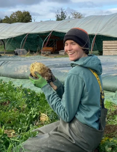 Lachende Gärtnerin kniet auf einem Feld und hält eine frisch geerntete Knollensellerie in die Kamera.