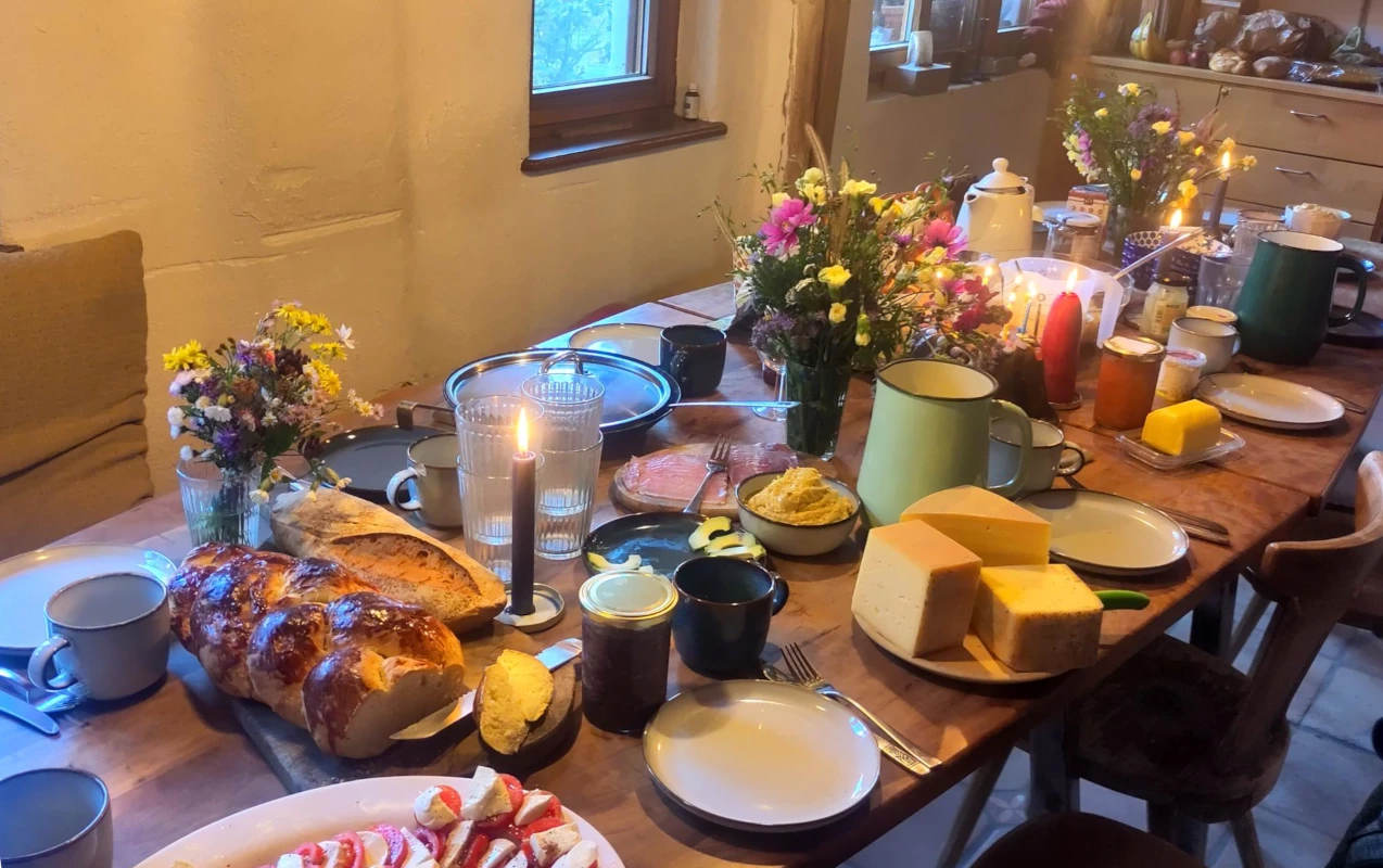Gemeinschaftliches Abendessen am gedeckten Tisch Reich gedeckter Holztisch in einer rustikalen Stube mit Kerzen, frischem Brot, Käse und Wildblumensträußen, der eine gemeinschaftliche Mahlzeit zeigt.
