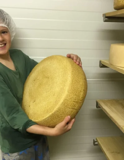 Fröhlicher Junge mit Haarnetz hält einen schweren, goldenen Käselaib in den Händen; Szene im Reifekeller der Solawi Luzernenhof vor Regalen mit Käse.
